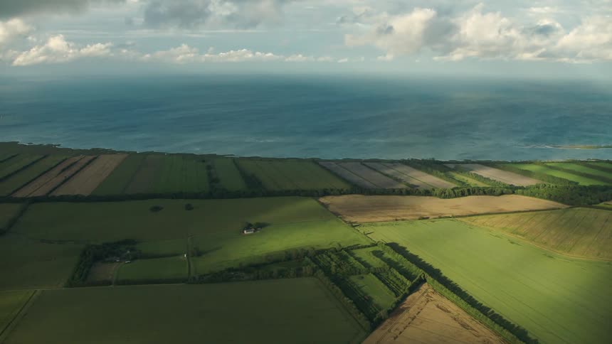 An aerial view of coastal farmland with the ocean and horizon in the background on a partly cloudy day