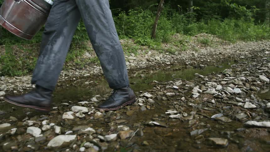 A man is walking while carrying a bucket along a rocky pathway in a woodland area during the day