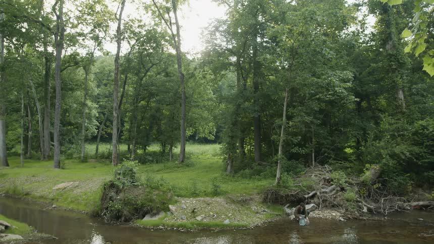 Man with brown shirt blue pants and black boots takes a bucket of water from river in the forest