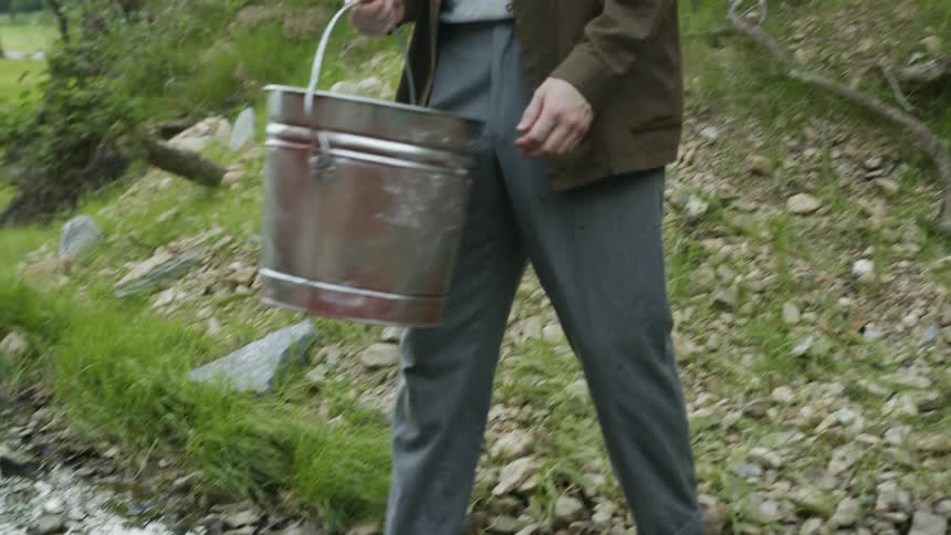 A man dipping a bucket into a river with the trees the sky and clouds in the background