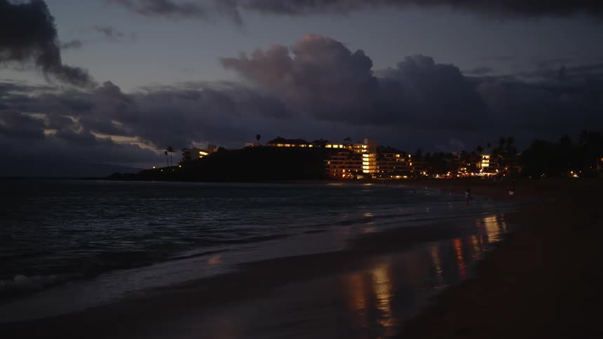 Person walks across beach at night as surf comes in with lights from buildings in background reflected by wet sand
