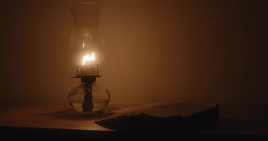 Flame flickers in lantern on wooden furniture near open book in dim shadowed room