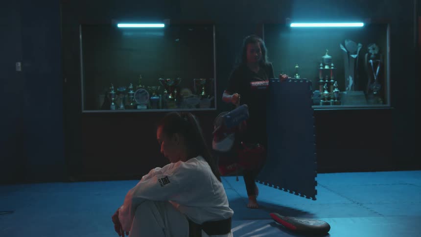 A woman in a gi sitting down on the floor in a room with a shelf trophies and a wall in the background