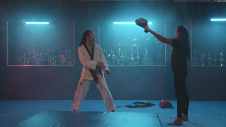 A woman is practicing martial arts with an instructor in a darkened room displaying many trophies and awards