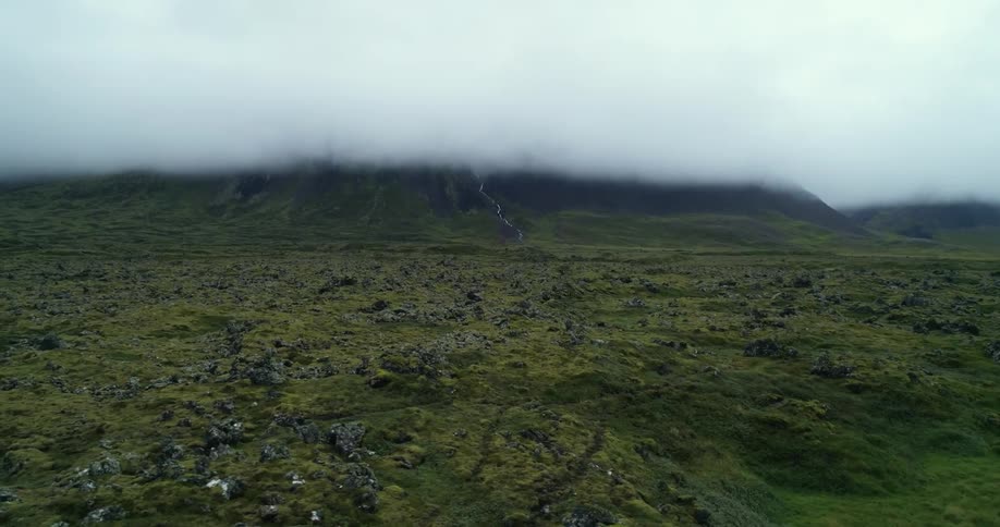 A stream of water flows down a green slope from under thick cloud cover