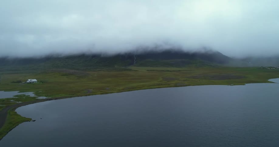 Road hugs water passing farms in countryside with mountain under low lying clouds in background