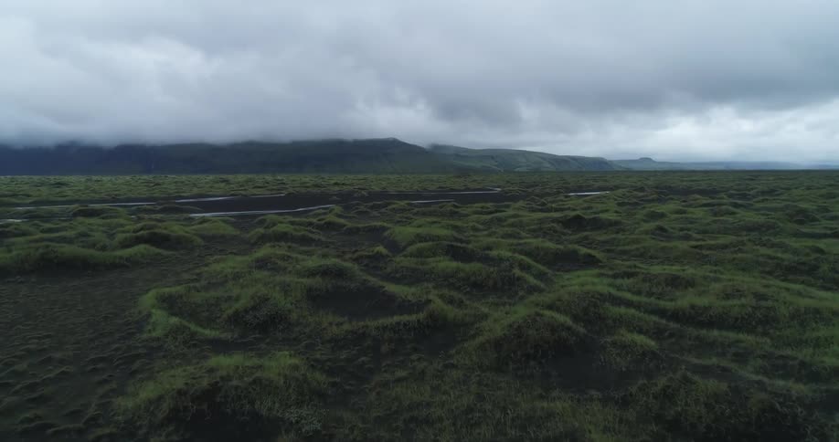 A stream of water curves and flows on a green bumpy terrain at the base of a mountain with heavy cloud cover