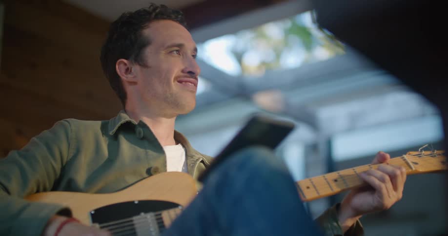 A man smiles as he looks at his phone and plays an acoustic guitar