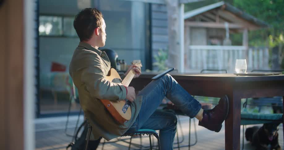 A man is sitting on a porch playing an electric guitar with his dog laying nearby during the day