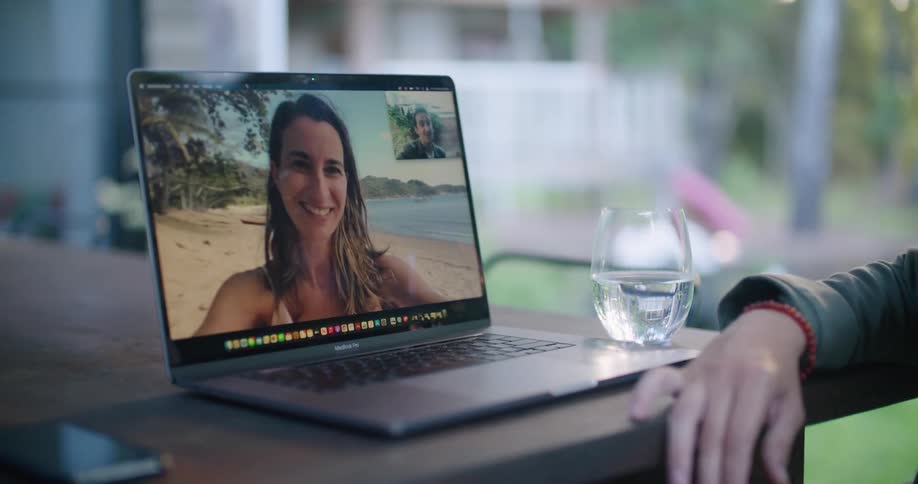 Man sitting at table in backyard talks with woman on beach via laptop they wave and kisses at each other
