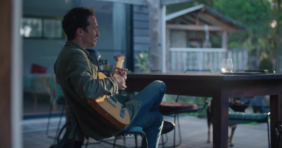A man is sitting on a porch in the daytime and playing an electric guitar and singing while a dog stands nearby wagging its tail