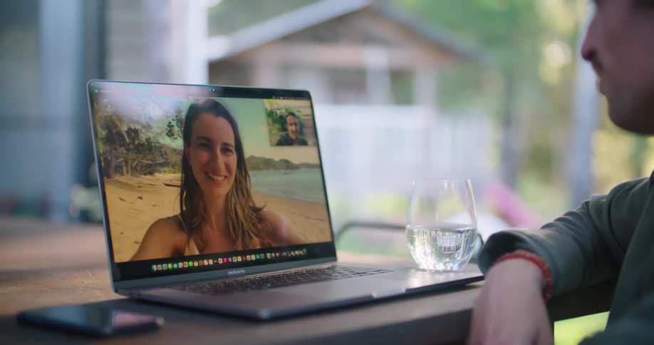 Man with green jacket and earring sits at table and makes a video call with a woman with brown wavy hair on the beach outdoors