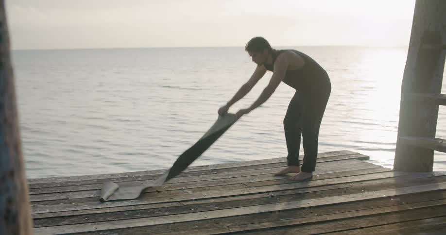 A man sets up a yoga mat on a wooden deck over the ocean