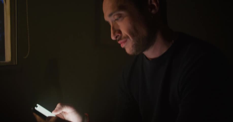 A man sitting inside a room is holding his phone in both hands and making a call