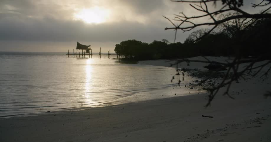 Water is gently rolling in onto a sandy beach near an old pier at sunset