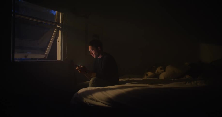 A man sits on the edge of his bed in the dark and texts on his smartphone