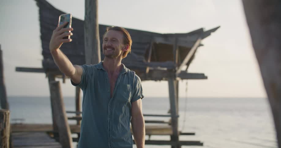 A man stands on the shore and holds his cell phone in front of his face and waves as he video chats