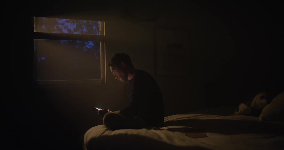 A man is sitting in the dark on a bed facing a window as he looks on his phone