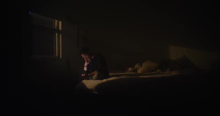 Man sitting on the bed in a dark bedroom looking at his phone