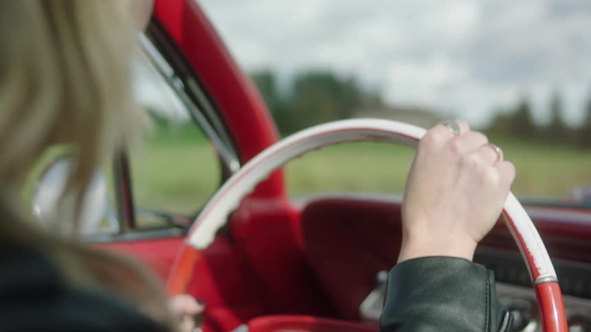 Blonde woman wearing sunglasses and black leather jacket driving a vintage red car on the countryside
