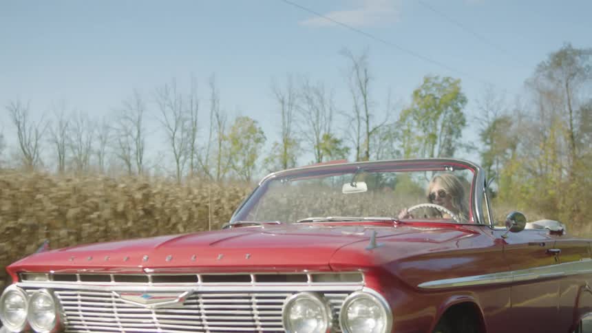 A woman is driving a classic car in a rural area with corn crops during the daytime