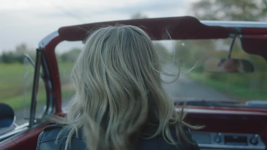Young blonde woman wearing sunglasses and black leather jacket driving a red vintage car in the countryside