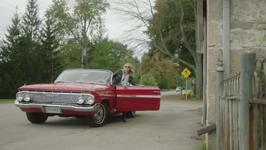 A woman is smoking a cigarette then gets into a red vintage convertible car and drives away