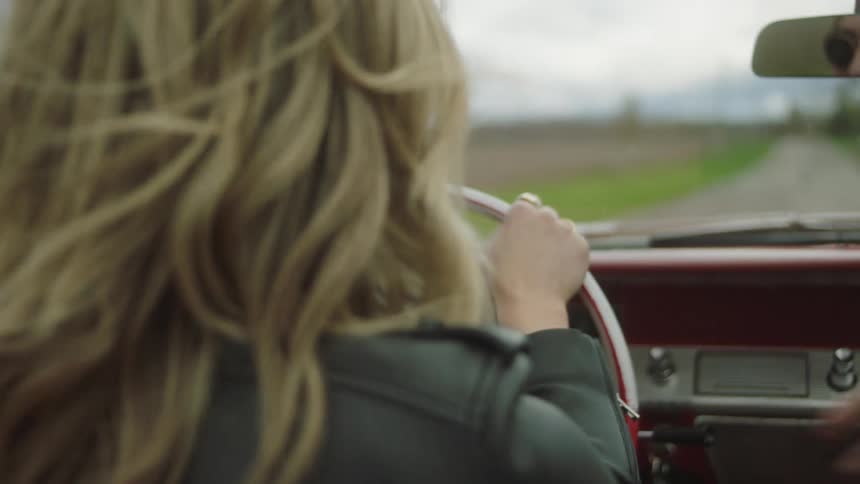 A woman raises her arm out of her convertible and laughs with a friend as they drive down a dirt road