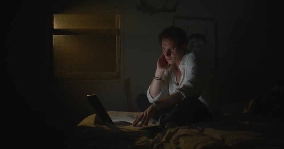 A man is at home typing on his laptop while sitting on a bed at night