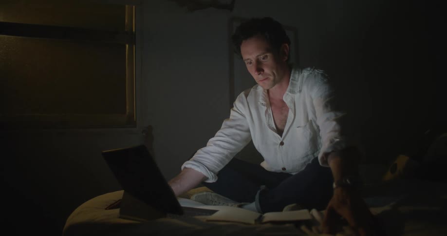 Man sits cross legged on bed typing on laptop with touchscreen