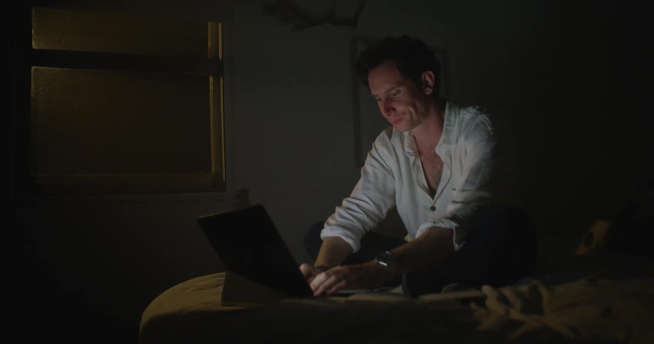 Man sits cross legged on bed in dark bedroom typing on laptop rubbing his face and checking the time