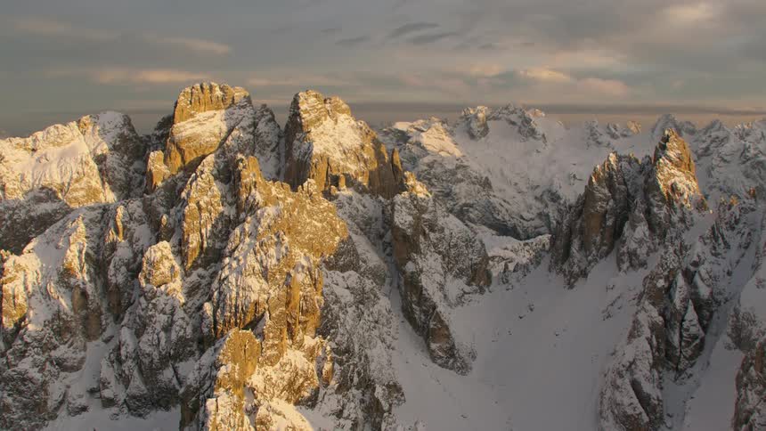An aerial view of a snowy mountain landscape with sunlight highlighting the peaks in the evening