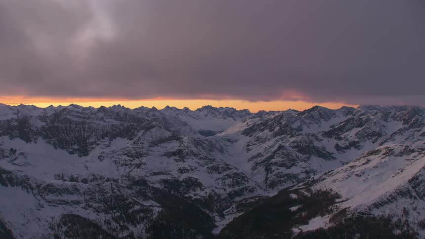 Thick clouds loom over a snowy mountain range at sunset