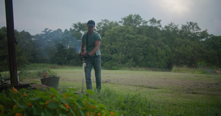 A man is using a string trimmer in a grassy yard on a cloudy day