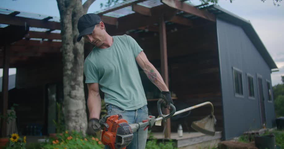 A man pulls the starter cord over a weed whacker while standing in the yard of a small house during the day