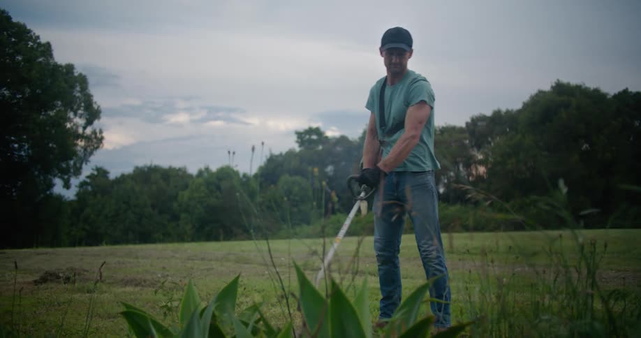 A man uses a string trimmer to cut grass in a field on a cloudy day