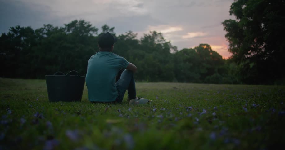 A man is sitting in the grass of a field surrounded by trees at dusk