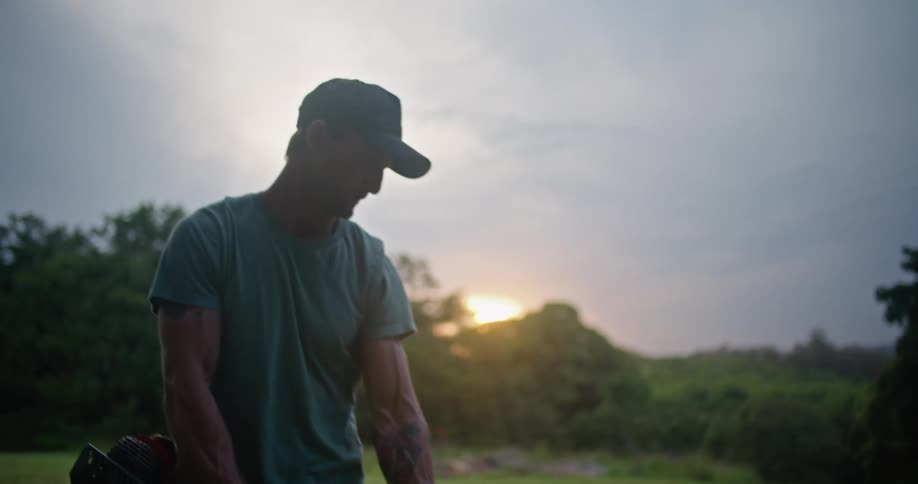 Man runs weed trimmer over grass in field with sun on horizon