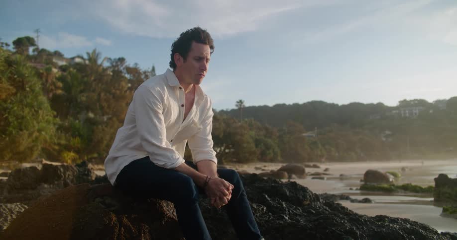 A man wearing a white shirt is sitting on rocks on a beach and looking around