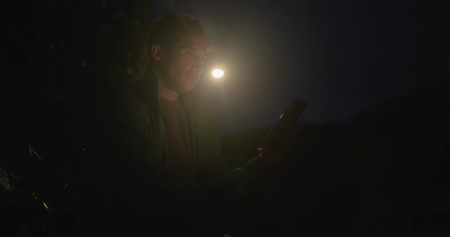 A man is outside at night texting on his cell phone as a bright light shines behind him