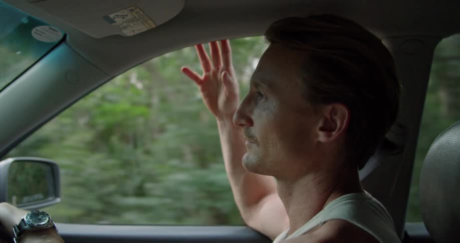 A man is driving on a road with his arm leaning out of the window and passing by many trees