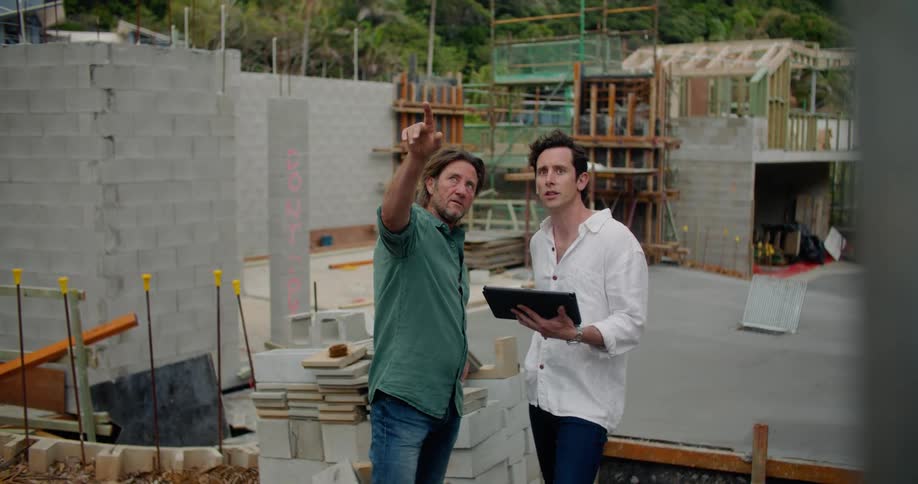 Two men are standing at a building construction site talking and pointing with an electronic tablet