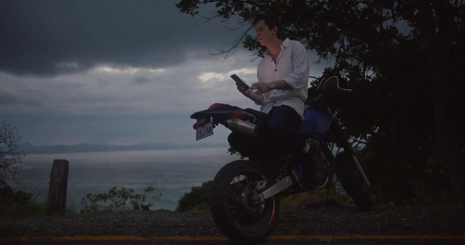 A man looking at a smartphone while sitting on a parked motorcycle on a beach at dusk