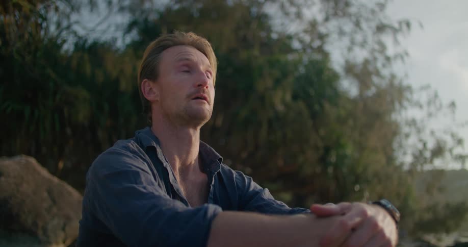 A man is sitting outside looking up at the sky and scrunching his eyes shut