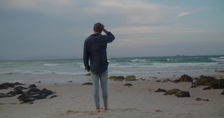 A man runs his hand through his hair as he stands on the beach and looks at the ocean