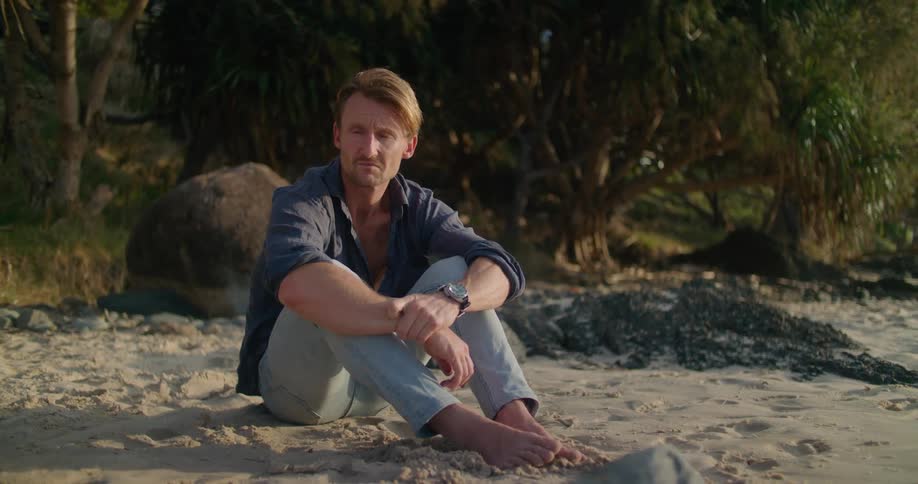 Blonde man with blue shirt black wristwatch and jeans sits on sand beach next to sea