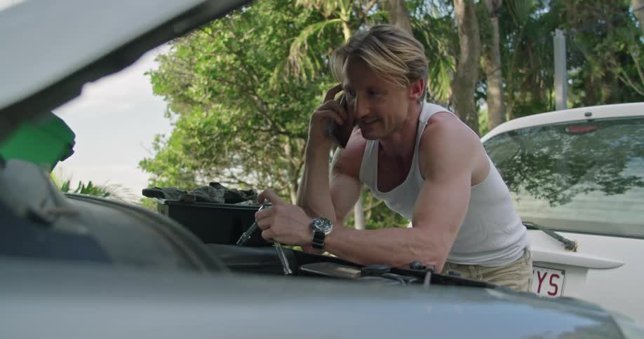 Blonde man with white undershirt and brown pants repairs car engine next to forest