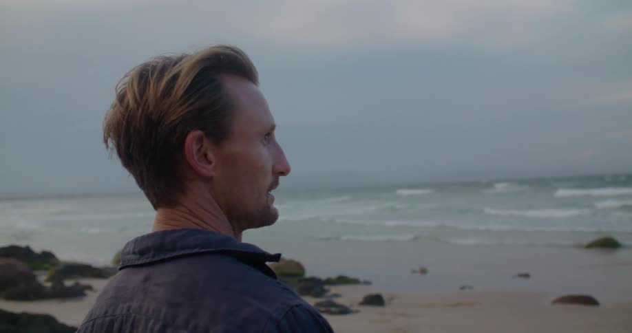 A man stands on the beach and looks out at the water and runs his hand through his hair and scratches the back of his head