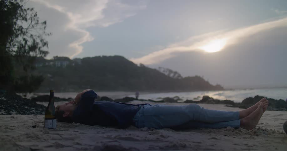Man with blue shirt and jeans lies down on the beach and smokes next to wine bottle at sunset