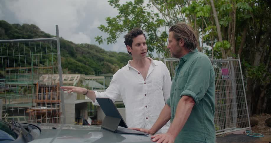 A man stands with hands on a car hood with a laptop open at a construction site listening to another